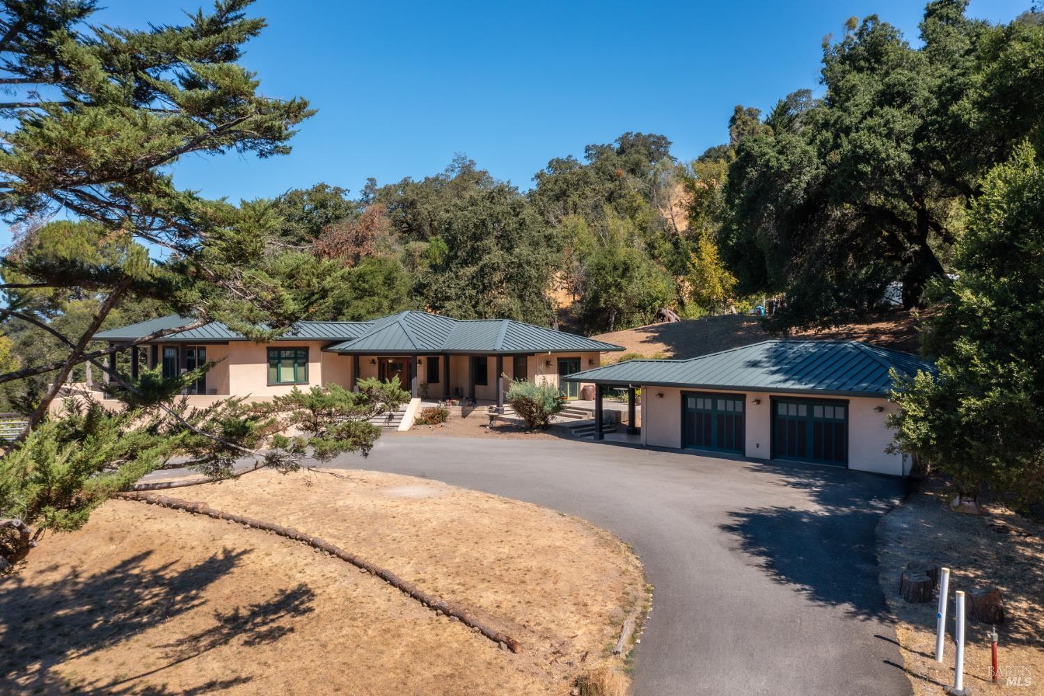 Single-story stucco home with green metal roofs and a two-car garage among mature trees.